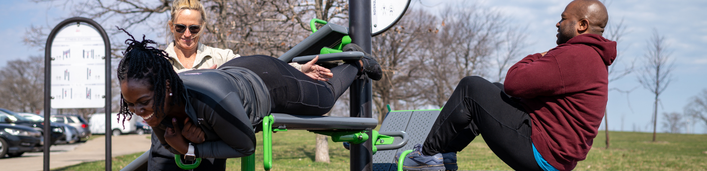 Three people exercising on outdoor gym equipment. One person does back extensions while another does sit-ups.